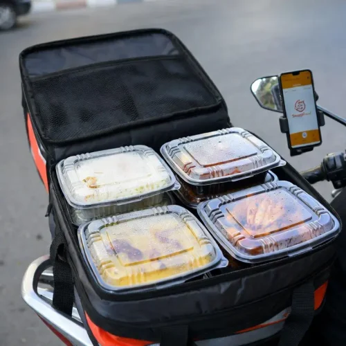 A professional, realistic wide shot of a cloud kitchen or dark kitchen facility serving multiple delivery platforms. Rows of stainless steel preparation tables show dozens of identical transparent plastic containers with secure lids being filled with food by kitchen staff in modest workwear (hijabs, aprons, gloves). A dedicated packing station has a label printer and thermal bags ready. On the wall, a large digital screen shows incoming orders from multiple platforms: "SnappFood: 12 orders", "Delino: 8 orders", "Local Market: 5 orders". Bright industrial LED lighting. Canon 5D Mark IV, 35mm lens, f/2.8, wide depth of field. Colors: silver tables, white and black uniforms, transparent containers, colorful food, blue digital screen. Iranian-Islamic context strictly respected – staff with proper hijab and modest attire. Square 1:1 ratio. Extremely realistic cloud kitchen photography.