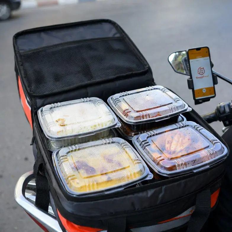 A professional, realistic wide shot of a cloud kitchen or dark kitchen facility serving multiple delivery platforms. Rows of stainless steel preparation tables show dozens of identical transparent plastic containers with secure lids being filled with food by kitchen staff in modest workwear (hijabs, aprons, gloves). A dedicated packing station has a label printer and thermal bags ready. On the wall, a large digital screen shows incoming orders from multiple platforms: "SnappFood: 12 orders", "Delino: 8 orders", "Local Market: 5 orders". Bright industrial LED lighting. Canon 5D Mark IV, 35mm lens, f/2.8, wide depth of field. Colors: silver tables, white and black uniforms, transparent containers, colorful food, blue digital screen. Iranian-Islamic context strictly respected – staff with proper hijab and modest attire. Square 1:1 ratio. Extremely realistic cloud kitchen photography.
